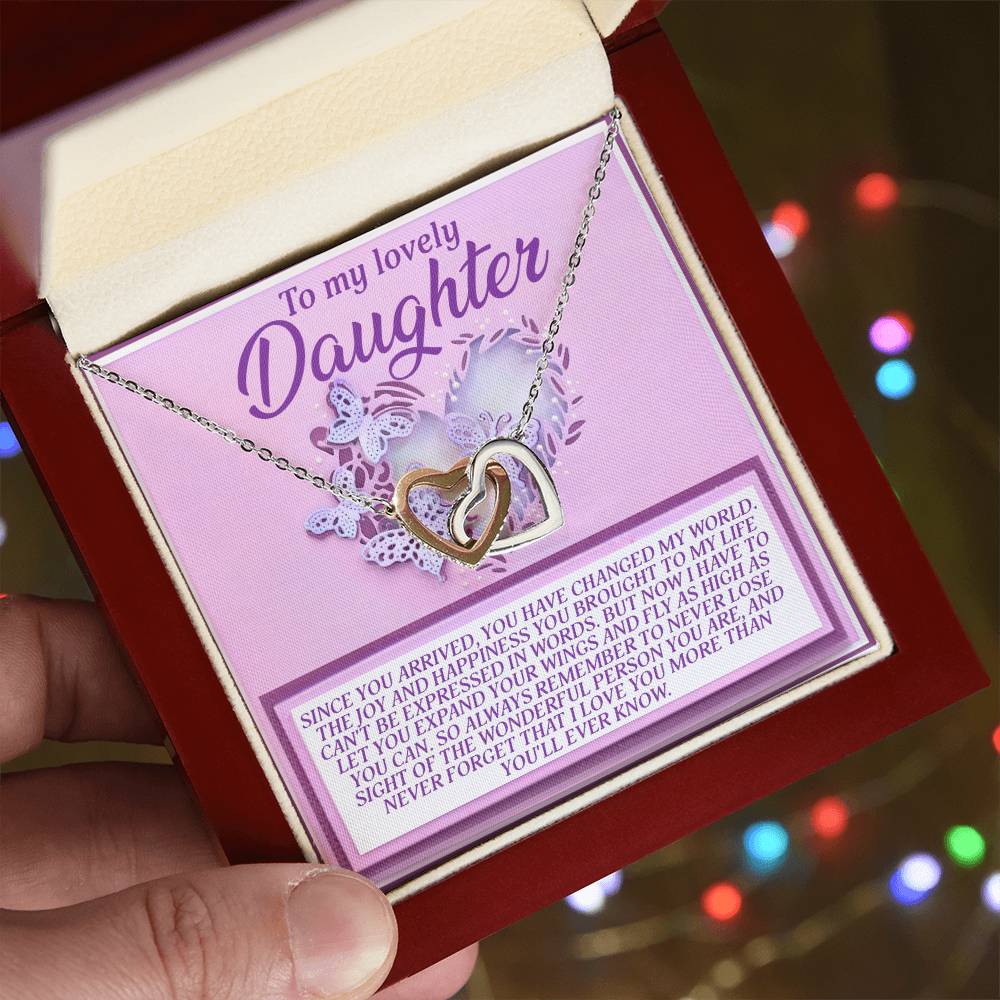 A hand displays an open jewelry box with the Daughter-Expand Wings Interlocking Hearts Necklace in rose gold. "To my lovely Daughter" is inscribed inside on a vibrant purple and pink background. Softly blurred, colorful lights twinkle in the backdrop.