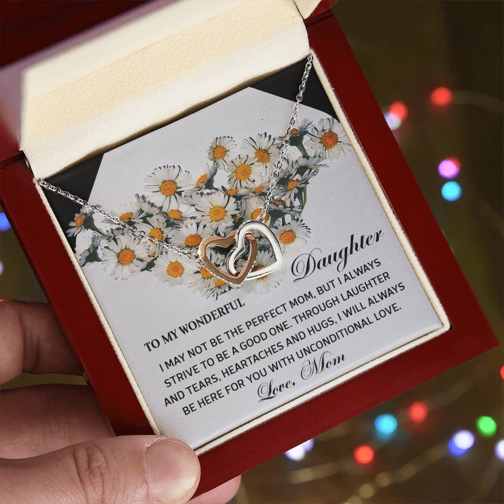 A hand unveils a jewelry box with the Daughter-Unconditional Love - Interlocking Hearts Necklace in surgical steel and rose gold, adorned with cubic zirconia crystals. A heartfelt message card for a daughter, bordered by illustrated daisies, nestles inside.