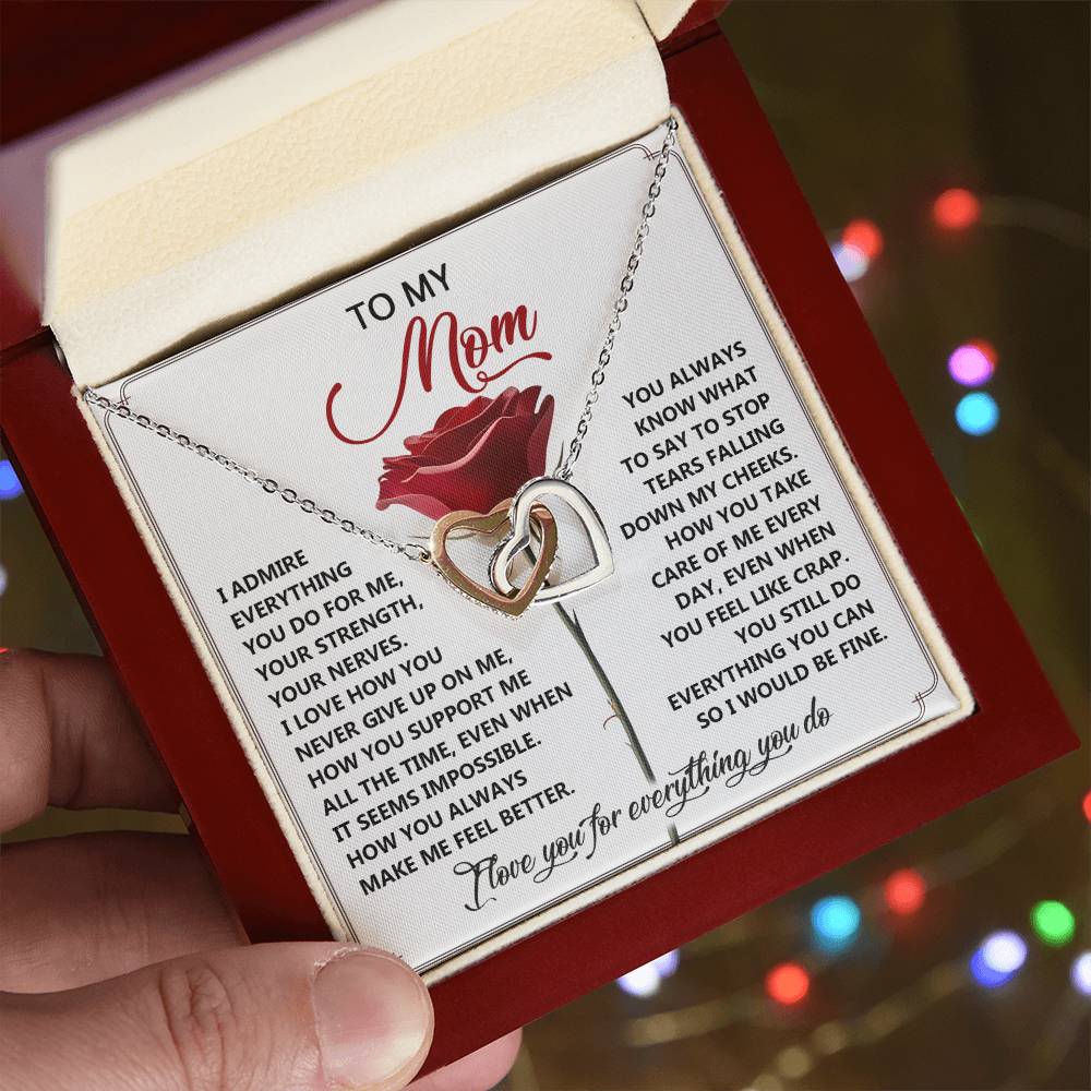 A hand holds an open red gift box with a card for a mom and the Mom-Be Fine - Interlocking Hearts Necklace featuring cubic zirconia crystals. The background is blurred with colorful bokeh lights, making it an ideal gift for her.