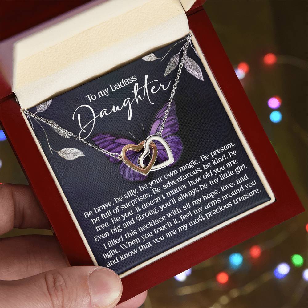 A hand presents an open box with the Daughter-Big And Strong - Interlocking Hearts Necklace adorned with cubic zirconia. Behind it, a card offers a heartfelt message to a daughter promoting bravery, kindness, and love. Shimmering colorful lights blur in the background.