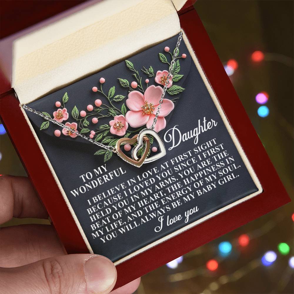 A person holds a Daughter-Love Sight box with an Interlocking Hearts necklace featuring pink flowers and cubic zirconia crystals. The lid has a loving message to a daughter, wishing her happiness. Blurred multicolored lights create a soft background ambiance.