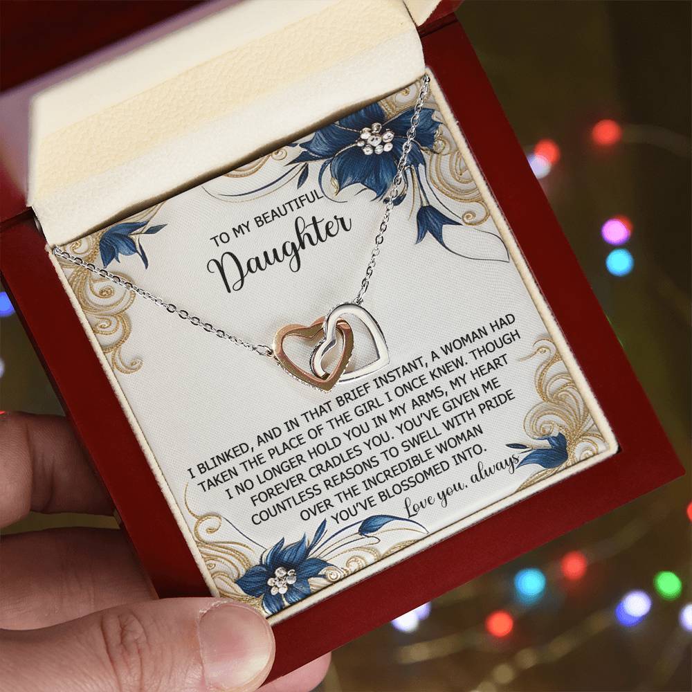 A hand unveils the Daughter-In My Arms - Interlocking Hearts Necklace, adorned with cubic zirconia crystals. A heartfelt message to a daughter appears on floral paper inside the lid. Blurred lights twinkle softly in the background.