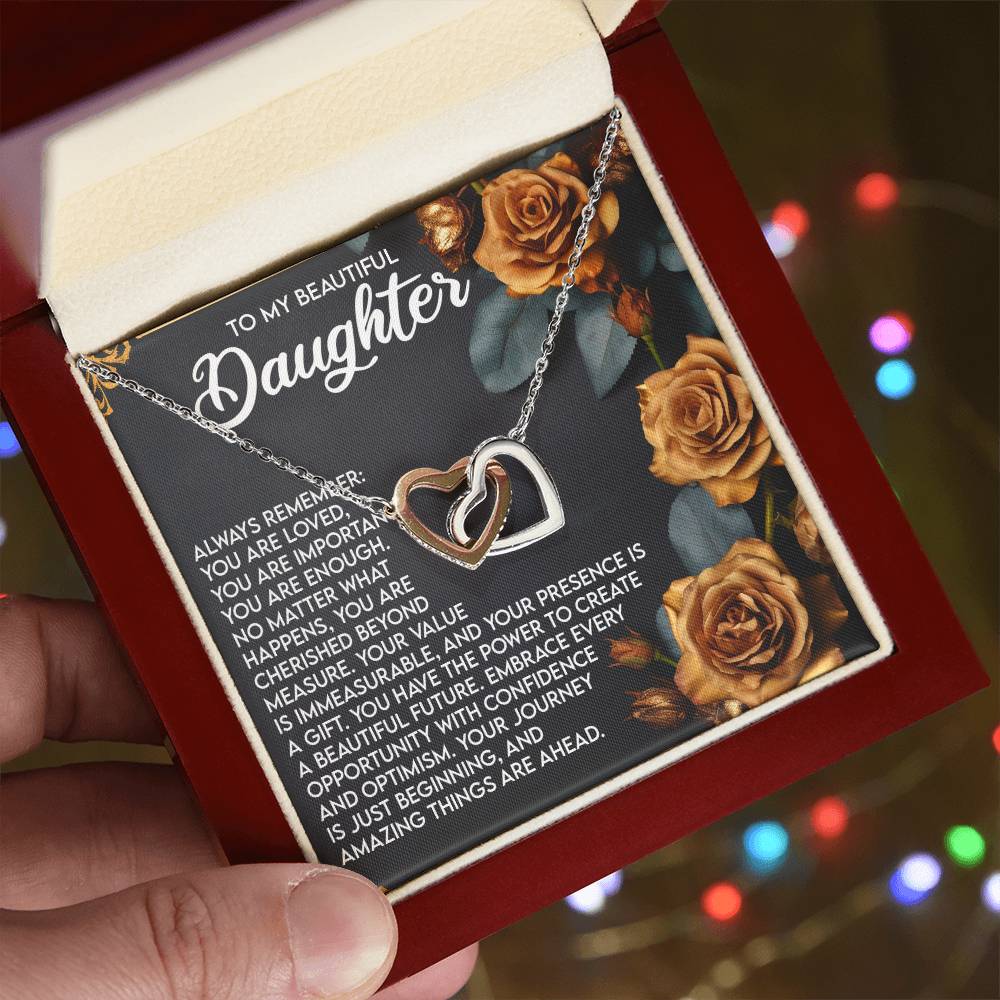 A person holds an open gift box revealing a stunning Daughter-Loved Enough - Interlocking Hearts Necklace, crafted with shimmering cubic zirconia crystals. Behind the necklace is a heartfelt message titled "To My Beautiful Daughter," adorned with golden roses, as festive lights softly blur in the background.
