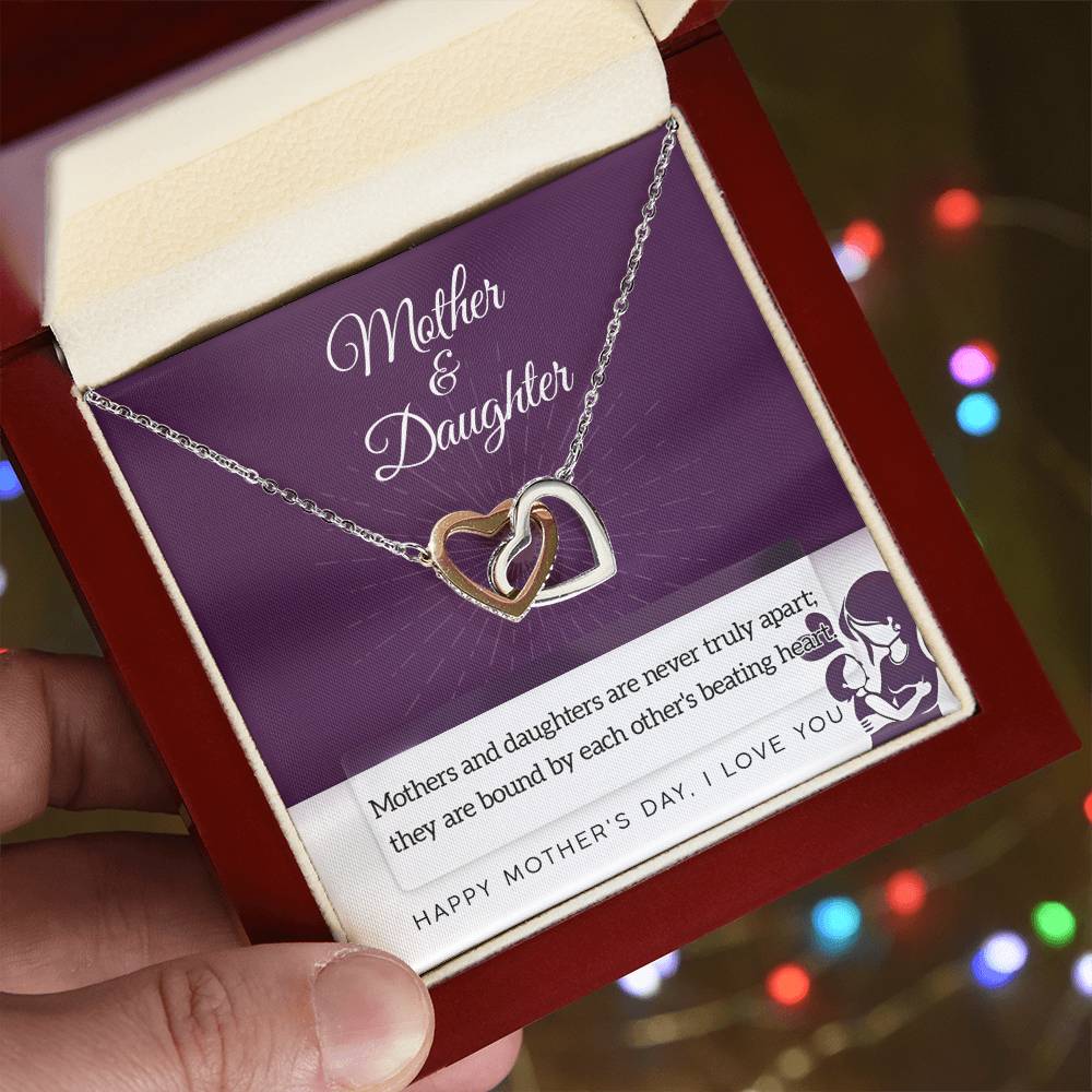 A hand holds an open jewelry box displaying the Mother & Daughter-Never Truly Apart - Interlocking Hearts Necklace with cubic zirconia crystals. Inside is a "Mother & Daughter" card with a heartfelt message, against colorful blurred lights.