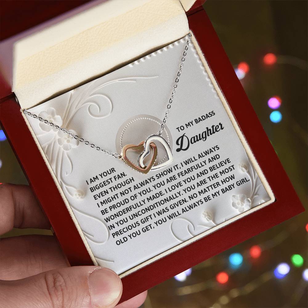 A hand presents a "Daughter-My Baby Girl" Interlocking Hearts necklace with rose gold details, housed in a box engraved with a heartfelt message. Multicolored lights twinkle in the blurred background, highlighting the radiant cubic zirconia crystals of the necklace.