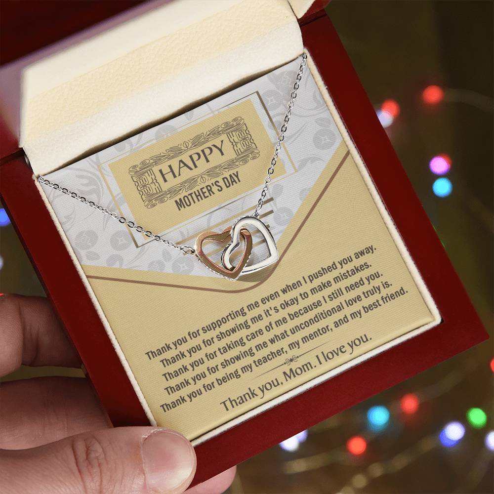 A hand holds an open red gift box revealing the Mother’s Day-Thank You - Interlocking Hearts Necklace with cubic zirconia crystals and a "Happy Mother's Day" card, with colorful blurred lights softly glowing in the background.