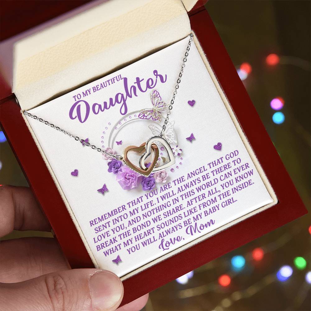 A hand holds an open jewelry box containing the Daughter-Angel Heart - Interlocking Hearts Necklace with cubic zirconia crystals. The box features a heartfelt message to a daughter from her mom, surrounded by purple flowers and heart decorations.