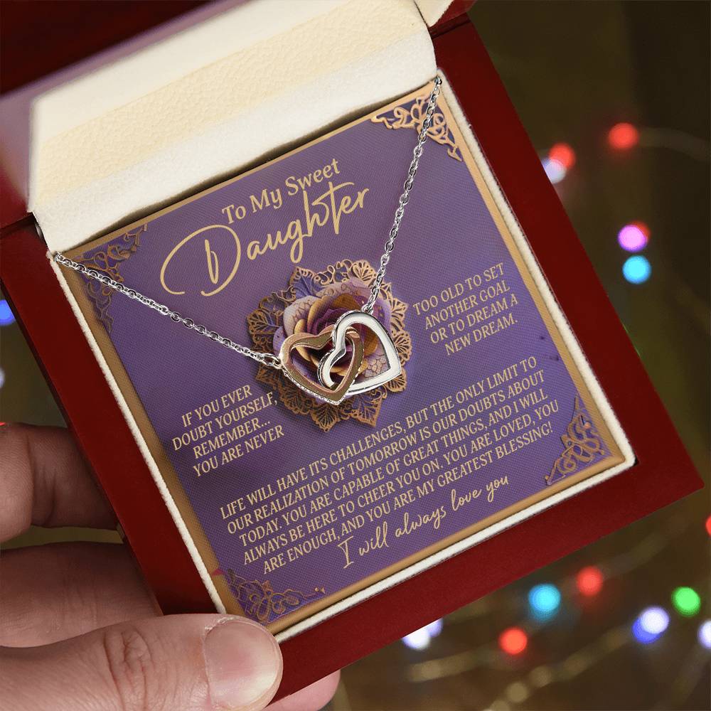 A hand holds an open red velvet jewelry box displaying the Daughter-Greatest Blessing - Interlocking Hearts Necklace with cubic zirconia crystals. A heartfelt message about dreaming and being loved is inside the lid, with blurred colorful lights in the background.
