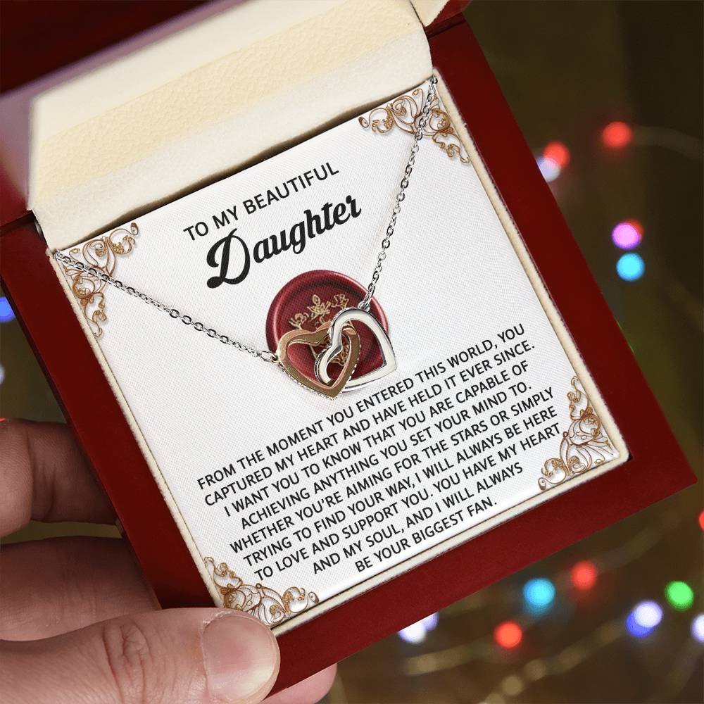 A hand holds an open jewelry box showing a silver interlocking hearts necklace adorned with cubic zirconia crystals. The lid features the heartfelt message "To My Beautiful Daughter" for the Daughter-Biggest Fan necklace.