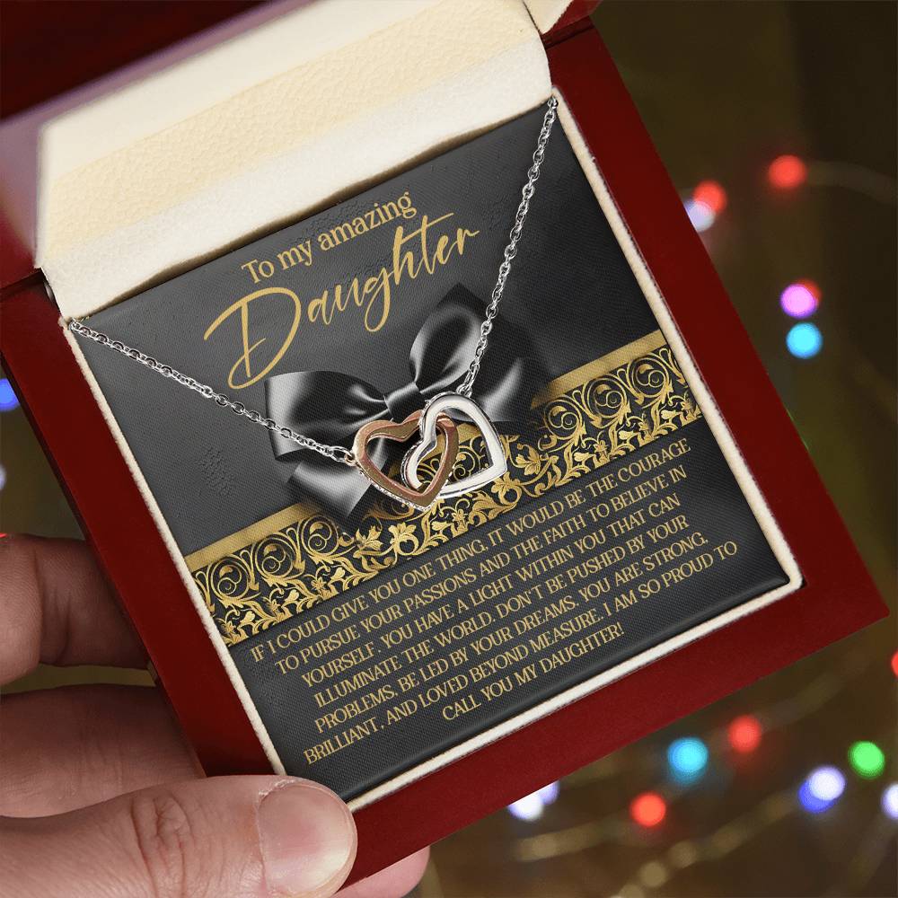An open jewelry box, held in hand, reveals the Daughter-Pursue Passions - Interlocking Hearts Necklace adorned with cubic zirconia crystals. A heartfelt message inside expresses pride and love for a daughter. The softly blurred background glows with colorful lights.