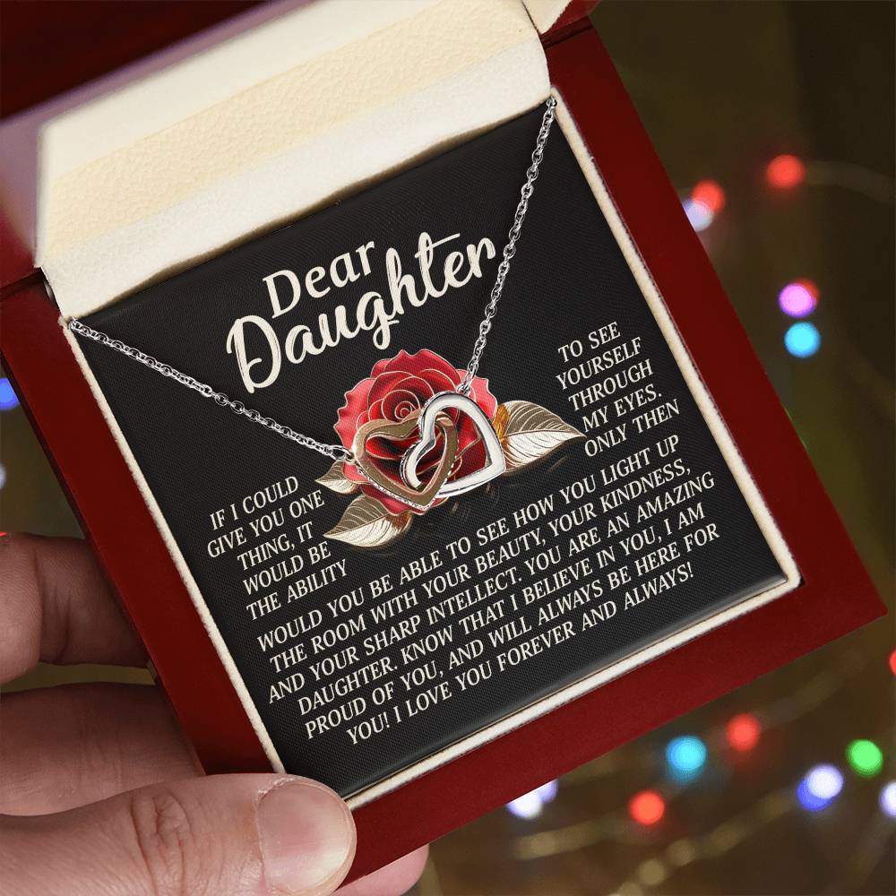 A hand presents the Daughter-Your Light - Interlocking Hearts Necklace, showcasing its rose design adorned with cubic zirconia. A heartfelt message to a daughter accompanies it. The background features blurred, twinkling colorful lights enhancing its rose gold finish.