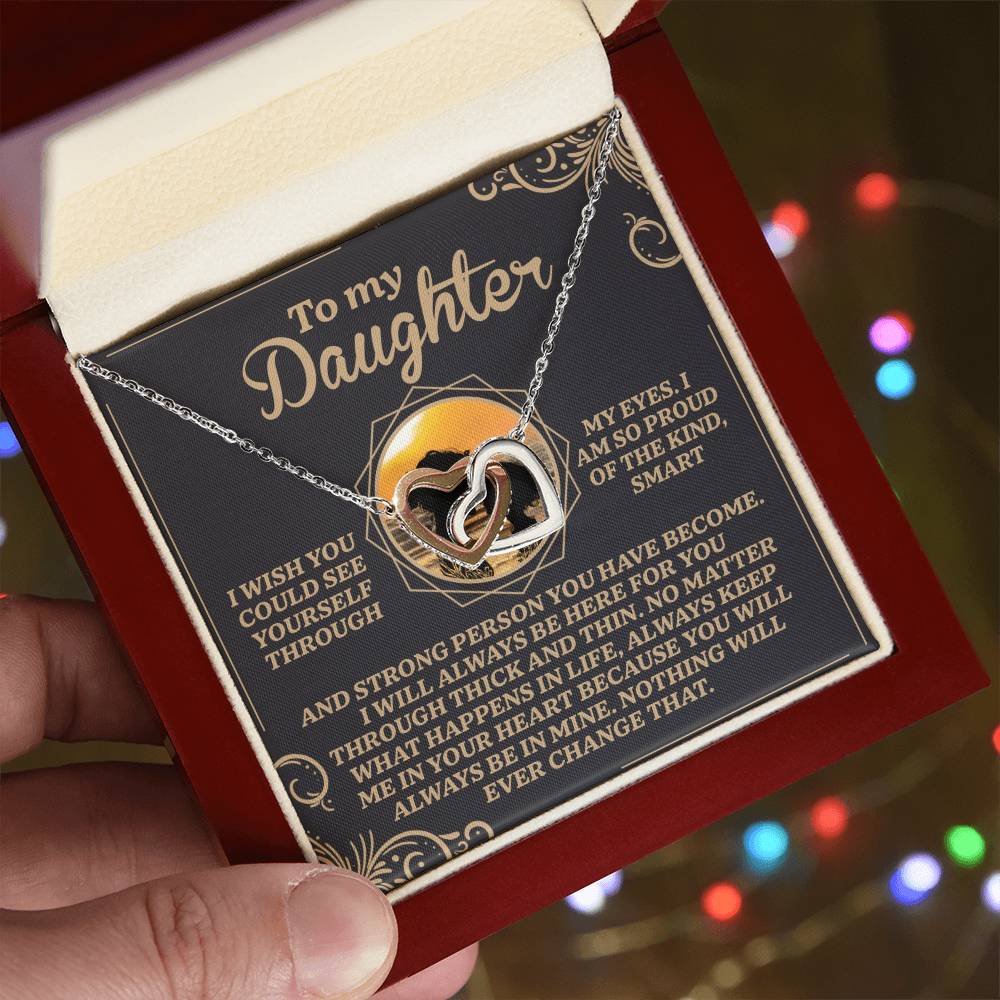 A hand holds an open jewelry box displaying the Daughter-Proud Heart - Interlocking Hearts Necklace with cubic zirconia crystals. Inside, a message conveys pride and love to a daughter, with soft colorful lights blurred in the background.