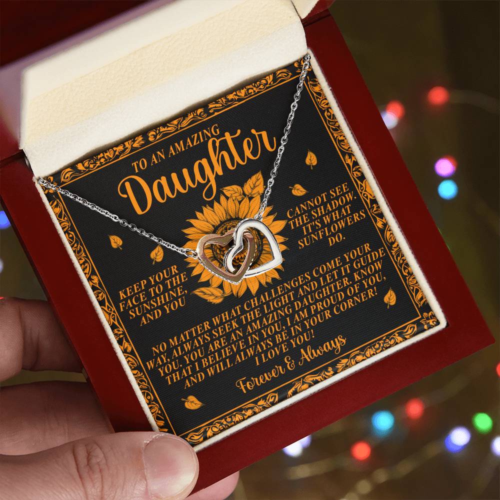 A hand holds an open jewelry box revealing the Daughter-Seek Light - Interlocking Hearts Necklace in a rose gold finish. Behind it, an ornate card reads "To an Amazing Daughter," set against a black and gold backdrop.