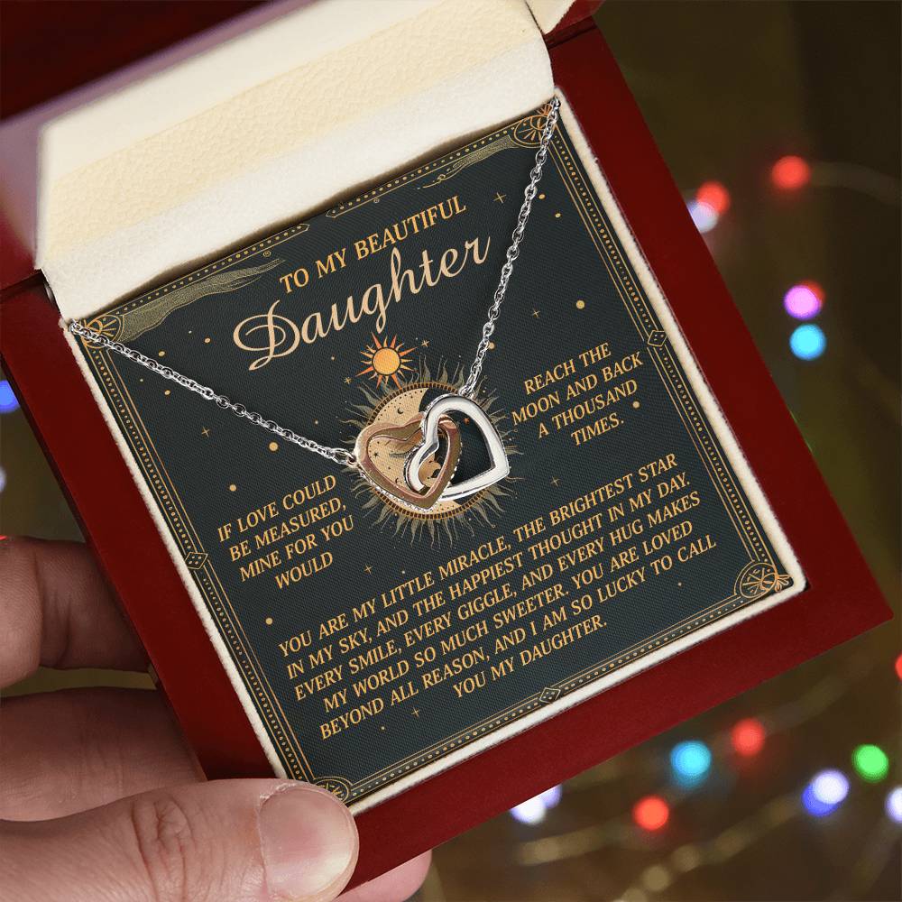 A hand presents an open gift box with the 'Daughter-To Call You' Interlocking Hearts Necklace featuring cubic zirconia crystals. The polished surgical steel box includes a loving message to a daughter, and multicolored blurred lights enhance the magical atmosphere.