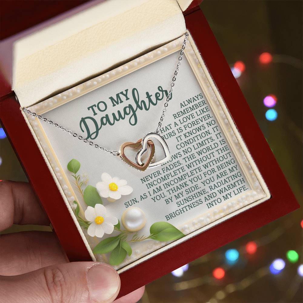 A hand presents a jewelry box displaying the Daughter-Love Forever - Interlocking Hearts Necklace in rose gold. The interior has a heartfelt message to a daughter amid a floral design with cubic zirconia crystals, set against a softly blurred background glowing with colorful lights.