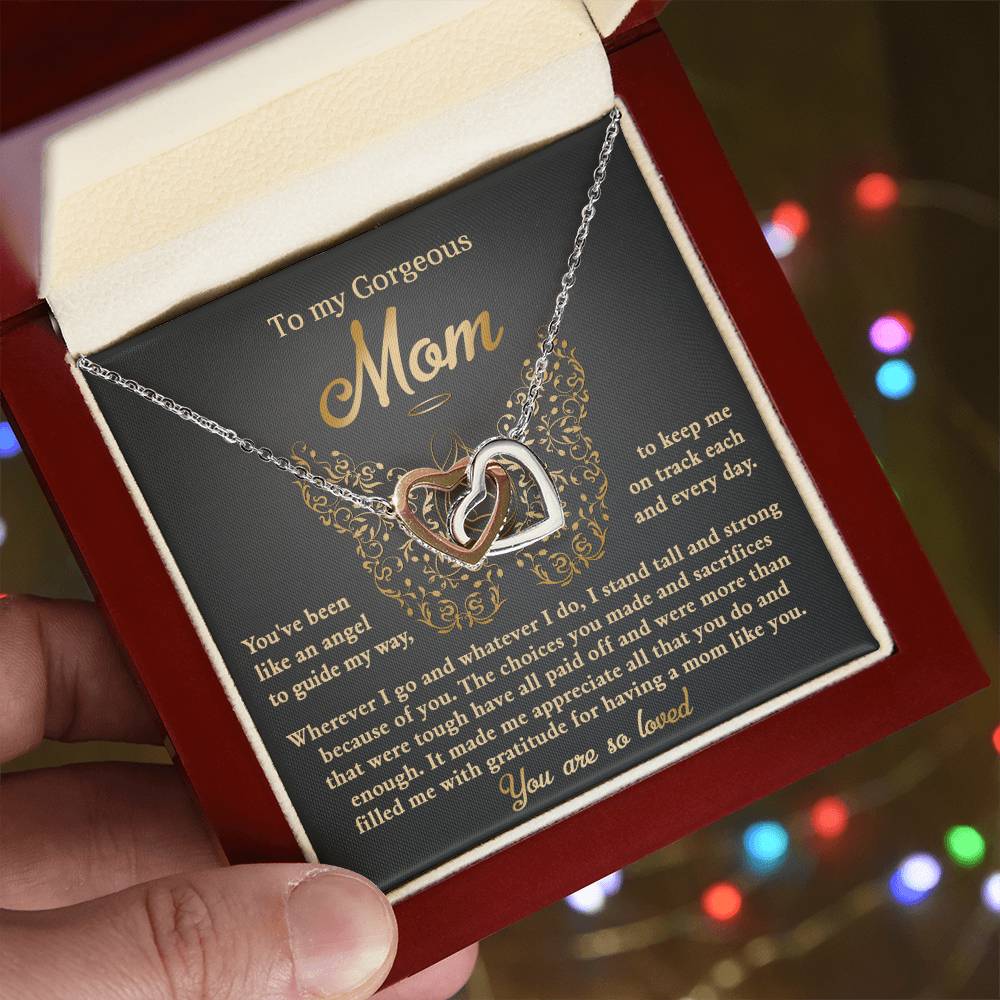 A hand holds an open jewelry box displaying the "To Mom, Like An Angel - Interlocking Hearts Necklace" adorned with cubic zirconia crystals and a card that reads "To my Gorgeous Mom" with an appreciation message. Softly blurred colorful lights enhance this unique gift's charm.