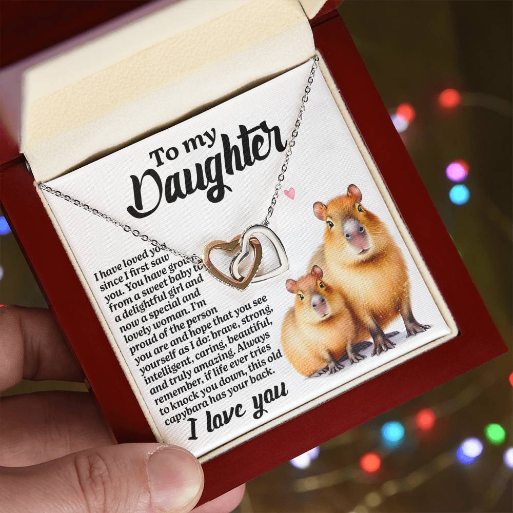 A person holds an open gift box revealing the Daughter-Sweet Baby - Interlocking Hearts Necklace adorned with cubic zirconia. The card reads "To my Daughter" and features two cute rodents, with blurred multicolored lights in the background.
