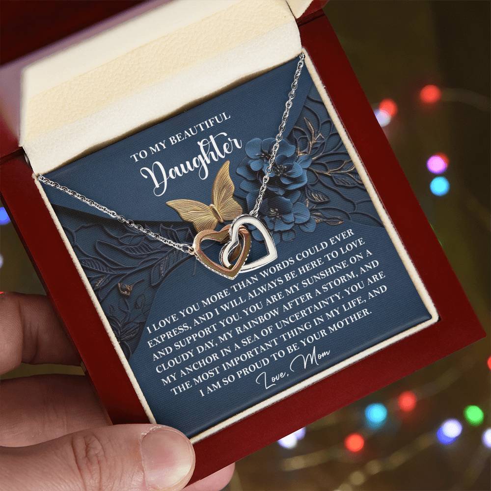 A hand gently cradles an open box revealing the "Daughter-In My Life - Interlocking Hearts Necklace," adorned with sparkling cubic zirconia crystals. Inside, a heartfelt message from a mother to her daughter speaks of enduring love and unwavering support.