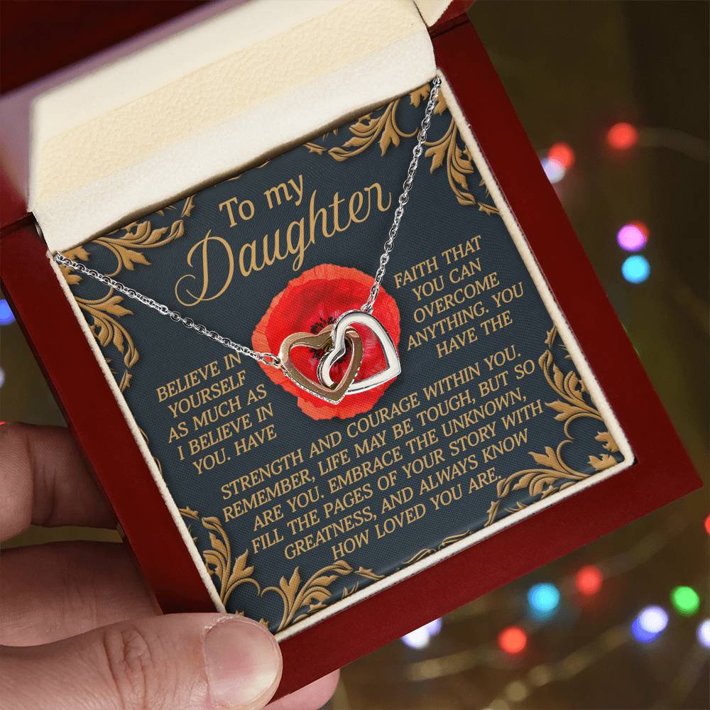 A hand holds an open jewelry box displaying the dazzling Daughter-With Greatness - Interlocking Hearts Necklace with a rose gold finish. The lid reads "To my Daughter" along with an inspirational message about faith, love, and strength. Cubic zirconia crystals shimmer against colorful blurred lights.