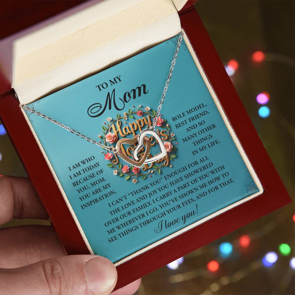 A hand holds the "Mom-Our Family - Interlocking Hearts Necklace" with a pendant spelling "Happy," featuring cubic zirconia crystals. The rose gold finish shimmers as a heartfelt note to "Mom" expresses gratitude and love amid soft, colorful lights.