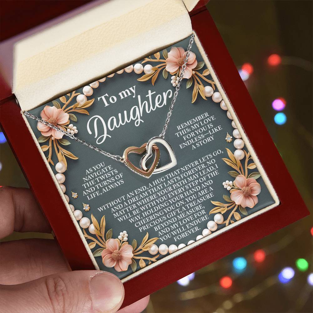 A hand holds an open jewelry box displaying the Daughter-Endless Love - Interlocking Hearts Necklace in rose gold, featuring pendants with cubic zirconia. A heartfelt message for a daughter is on the lid amidst floral and pearl designs. Colorful lights blur in the background.
