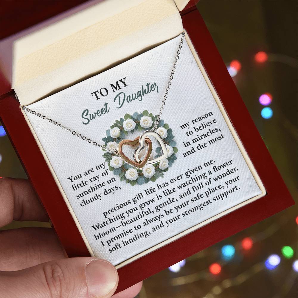 A hand displays an open red box with the Daughter-Strongest Support - Interlocking Hearts Necklace in a rose gold finish, accompanied by a card that reads, "To My Sweet Daughter," set against a blurred background of colorful lights.