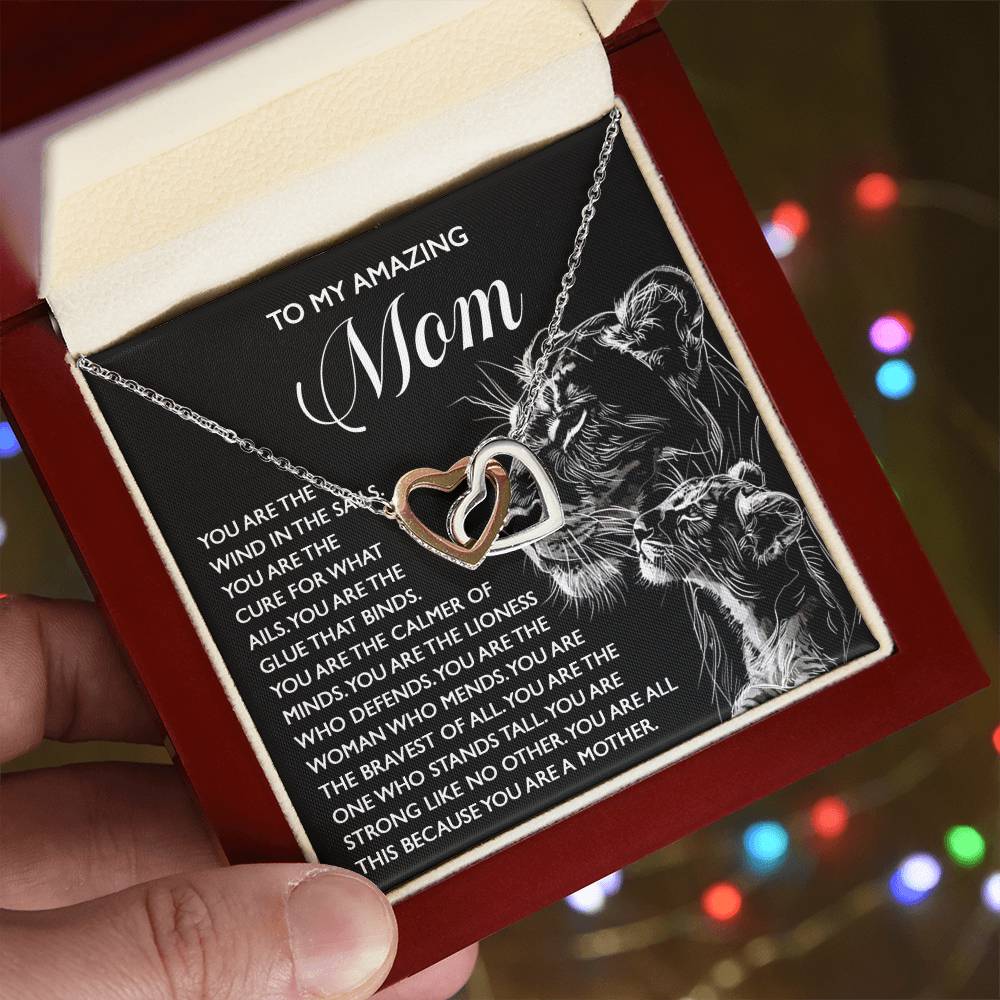 A hand displays the Mom-The Cure - Interlocking Hearts Necklace in a red jewelry box. The inner lid has a message to mom with a lioness and cub illustration, while soft bokeh from multi-colored lights enhances the scene.