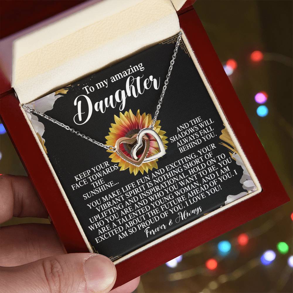 A hand holds an open jewelry box displaying the Daughter-Vibrant Spirit - Interlocking Hearts Necklace in a rose gold finish. Behind it, a black card with a heartfelt message to a daughter features a sunflower design, set against colorful blurred lights.