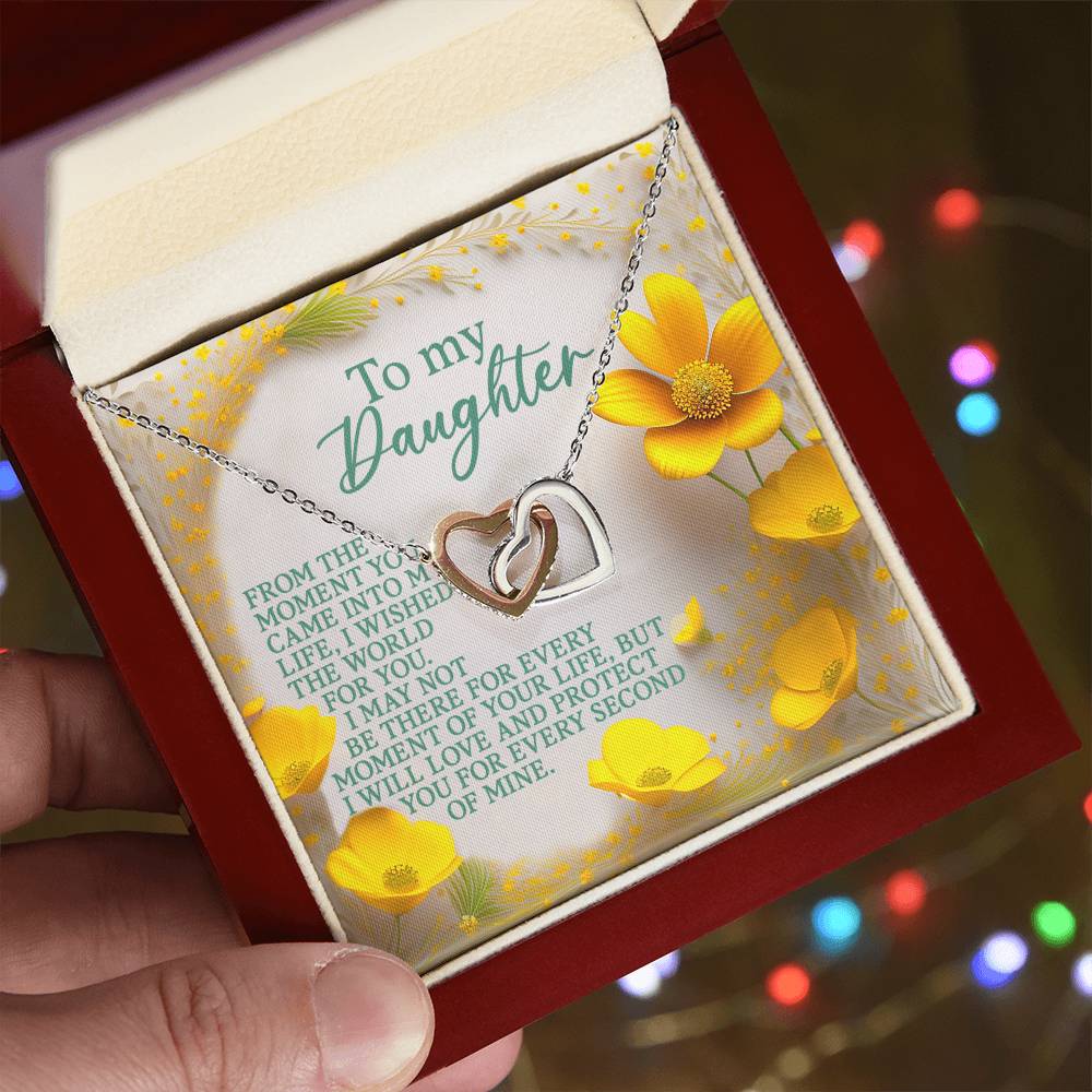 Someone holds a jewelry box containing the Daughter-Every Moment - Interlocking Hearts Necklace in rose gold. Inside, it reads "To my Daughter" with a heartfelt message and yellow flowers, set against softly blurred colorful lights highlighting its cubic zirconia crystals.