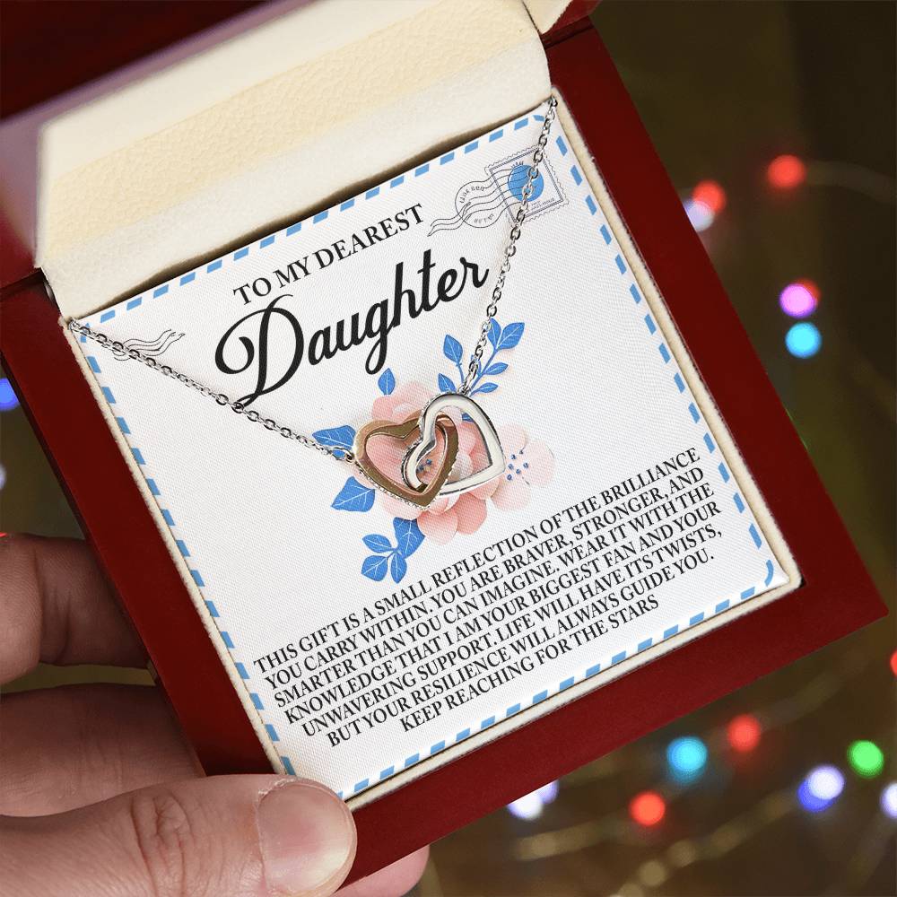 A hand presents a red jewelry box with the Daughter-Brilliance Within - Interlocking Hearts Necklace in rose gold, adorned with cubic zirconia. A heartfelt message for a daughter is shown against blue and pink floral designs, with blurred colorful lights twinkling in the background.