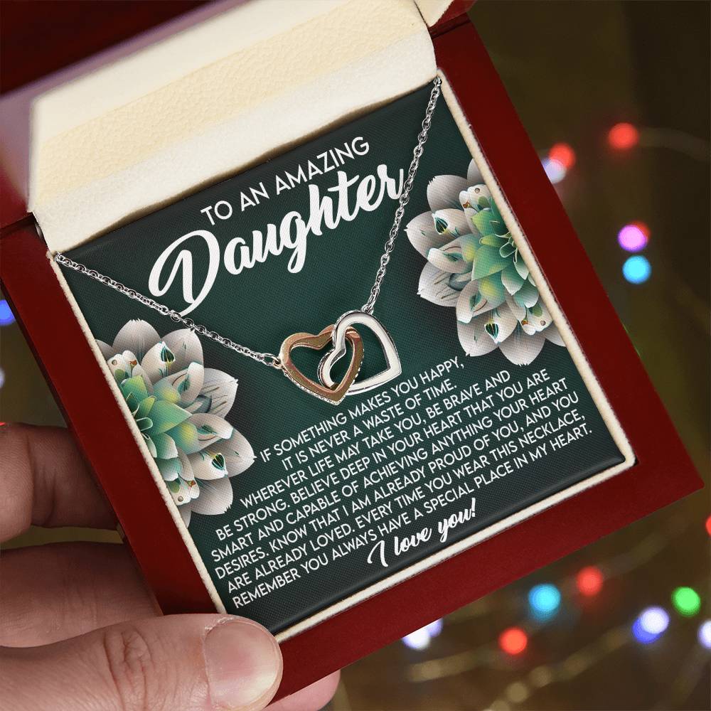 A hand holds an open jewelry box displaying the Daughter-Special Place - Interlocking Hearts Necklace in rose gold, embellished with cubic zirconia crystals. Behind it, a touching message to a daughter is framed by floral accents, with blurred multicolored lights in the background.