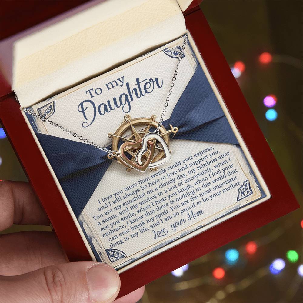 A person holds an open jewelry box with a Daughter-Sunshine Anchor - Interlocking Hearts Necklace featuring cubic zirconia crystals. Inside, a heartfelt message from mom to daughter expresses love and guidance, while softly blurred multicolored lights glow in the background.