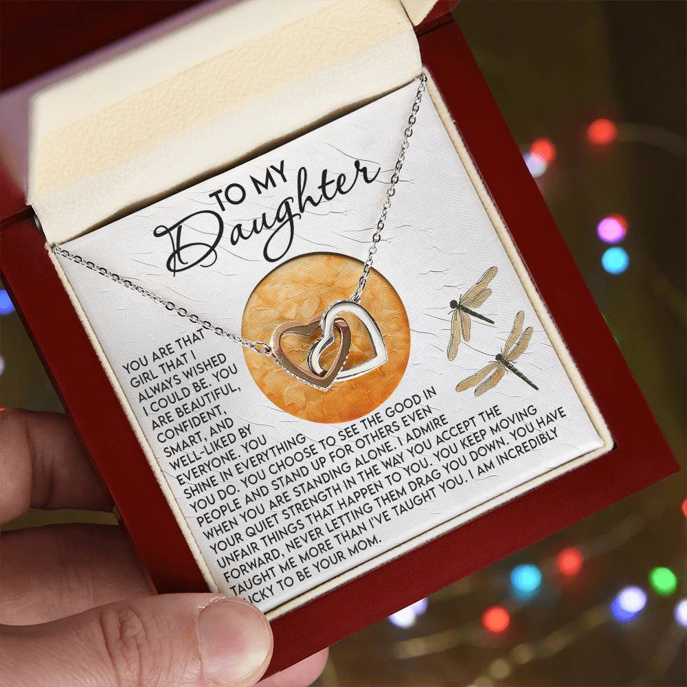 A hand holds an open jewelry box displaying the Daughter-Quiet Strength - Interlocking Hearts Necklace, a silver heart adorned with shimmering cubic zirconia crystals. Inside the lid is a heartfelt message to a daughter set against a blurred background with colorful lights.