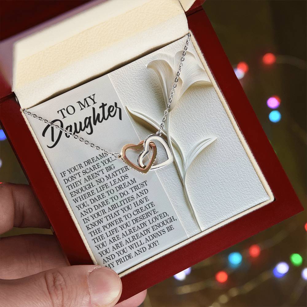 A hand holds a rose gold finish box containing the Daughter-Dare Dream - Interlocking Hearts Necklace adorned with cubic zirconia crystals. Behind it, a card reads "To My Daughter" with an inspirational message, set against a backdrop of blurred colorful lights.