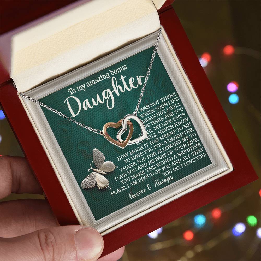 A hand holds a jewelry box with the "Daughter-Your Life - Interlocking Hearts Necklace" adorned with cubic zirconia. A butterfly charm adds grace, and the lid reveals a heartfelt message on a green backdrop, while festive lights twinkle softly in the background.