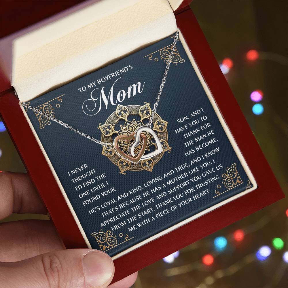 A hand holds an open jewelry box with the Mom-Thankful Heart - Interlocking Hearts Necklace, rose gold and cubic zirconia adorned. In the background, a heartfelt note to a boyfriend's mother expresses gratitude and admiration amid softly blurred twinkling lights.