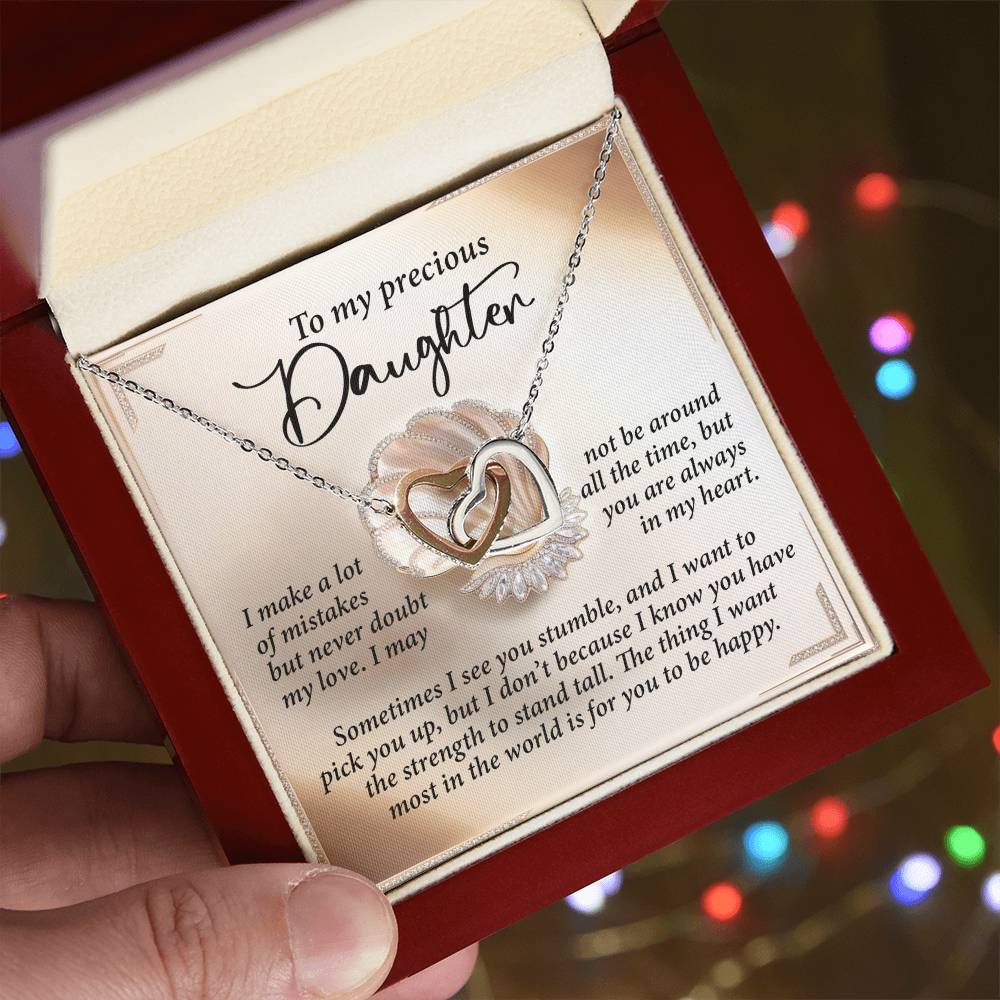 A hand holds an open jewelry box displaying the Daughter-Be Happy - Interlocking Hearts Necklace with a heart-shaped pendant. The 18k yellow gold finish shimmers with cubic zirconia crystals, and the box lid bears a sentimental message for a daughter as blurred multicolored lights twinkle behind.