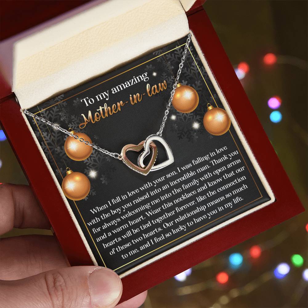 A hand holds an open jewelry box, revealing the Mother-in-Law-Two Hearts - Interlocking Hearts Necklace with a rose gold finish. The lid displays a heartfelt message to a mother-in-law, while multicolored lights blur softly in the background, adding a touch of sparkle reminiscent of cubic zirconia crystals.