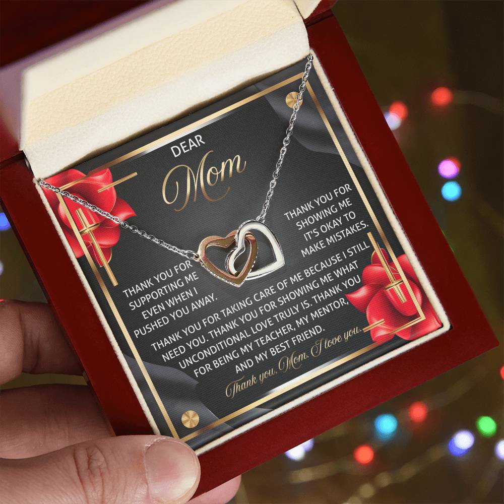 A hand holds an open red jewelry box with the stunning Mom-Best Friend 2 - Interlocking Hearts Necklace in a rose gold finish. The box shares a message of gratitude for "Mom" and her unwavering support, while colorful lights shimmer softly in the blurred background.