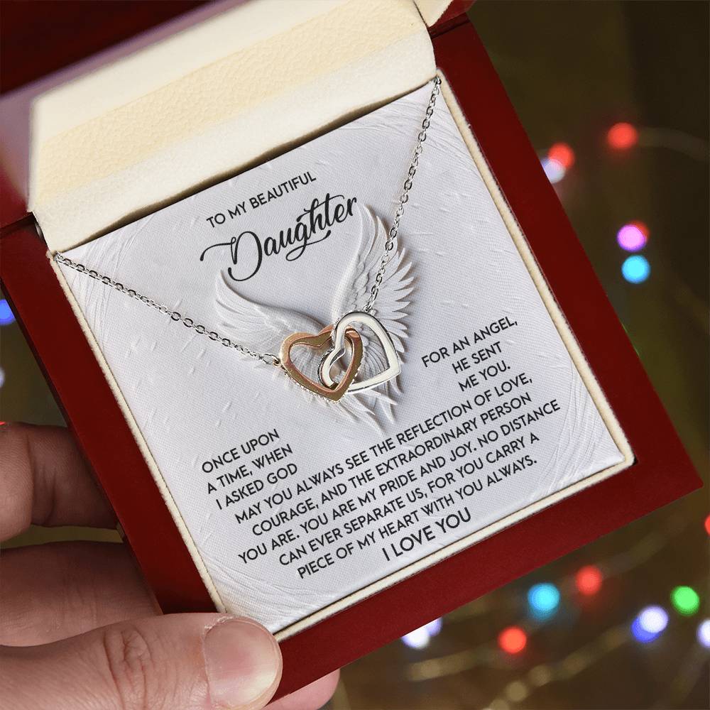 A hand displays an open jewelry box with the Daughter-An Angel - Interlocking Hearts Necklace, featuring cubic zirconia crystals. The lid expresses love and pride, affirming she's a piece of their heart, while the rose gold finish shimmers against colorful, blurred lights in the background.