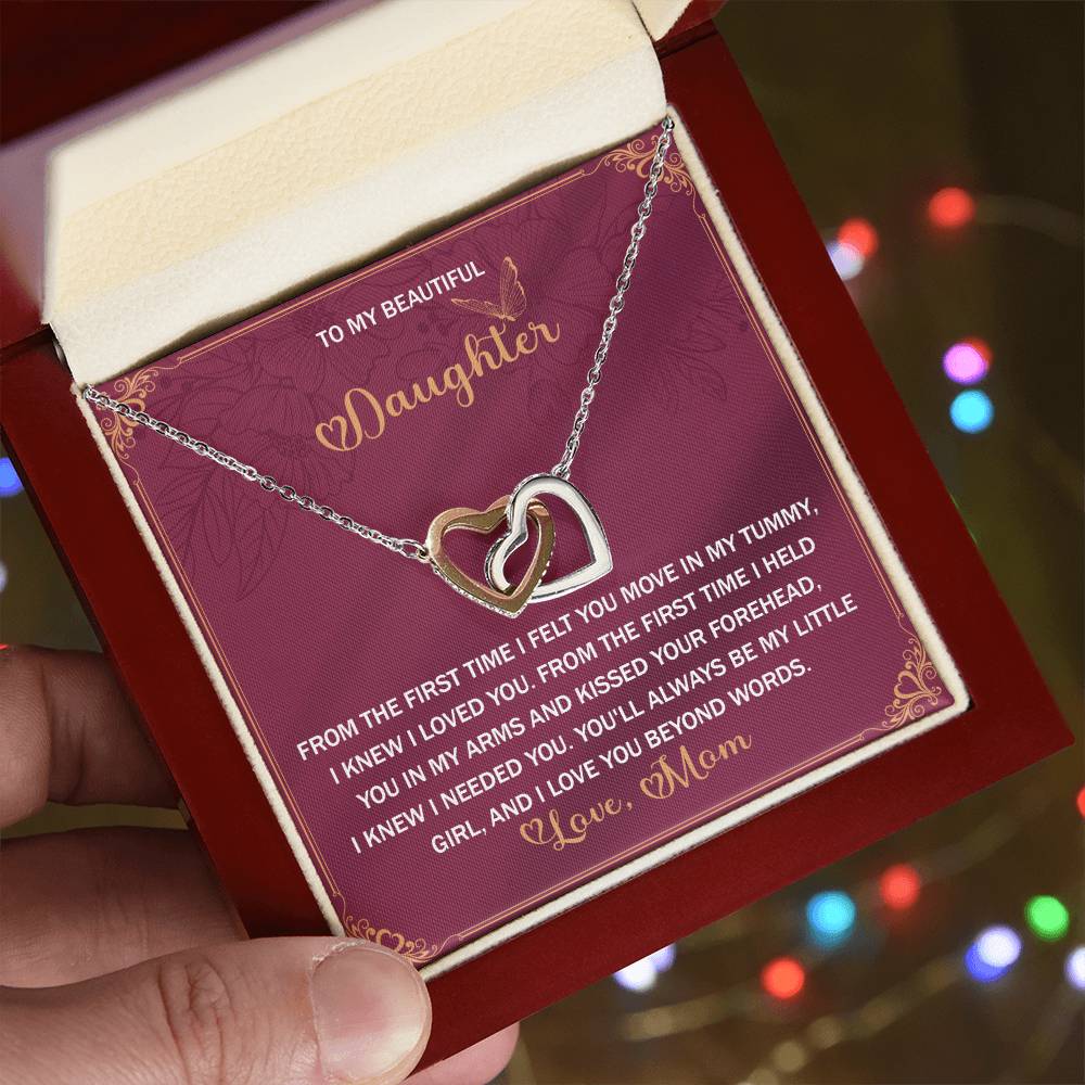 A hand offers a Daughter-Beyond Words - Interlocking Hearts Necklace in an open red jewelry box, showcasing a silver heart-shaped design with cubic zirconia crystals. Behind it, a card from "Mom" to "Daughter" expresses love and elegance.
