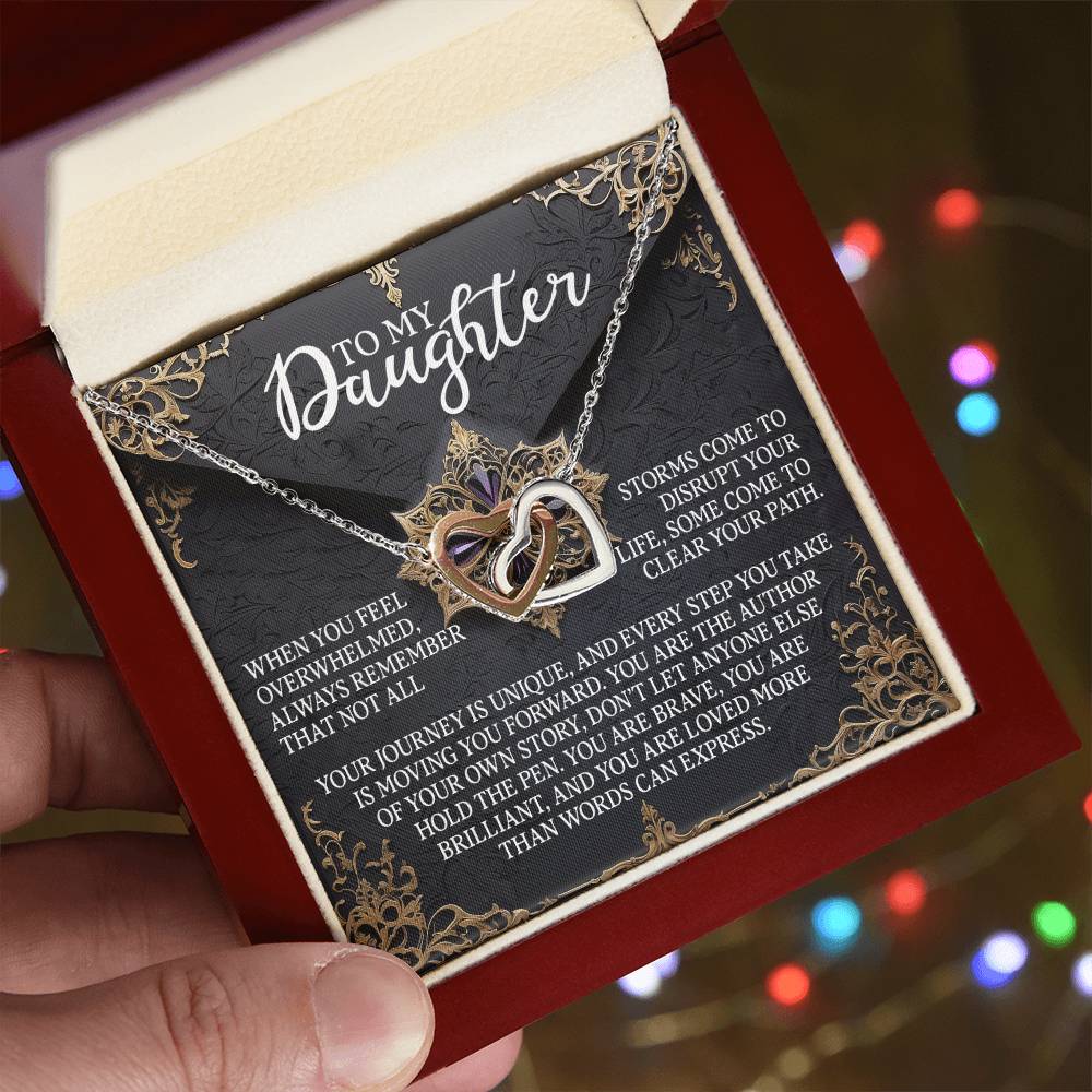 A hand holds an open red box with the Daughter-The Pen - Interlocking Hearts Necklace, featuring a rose gold finish. The lid reads "To My Daughter," and inside, a heartfelt message is in gold and white on a black background with ornate golden borders.