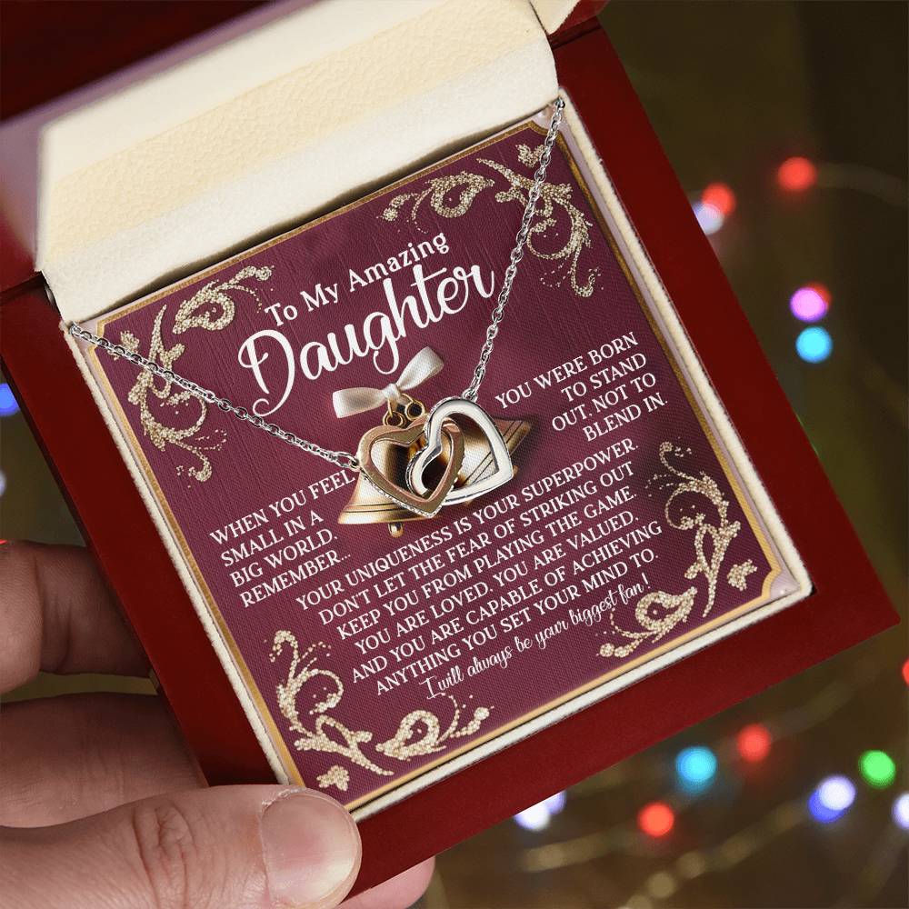 A person holds an open red gift box with the Daughter-Stand Out - Interlocking Hearts Necklace in rose gold finish. The card, adorned with decorative gold patterns, expresses a heartfelt message to a daughter. Blurred colorful lights form the background.