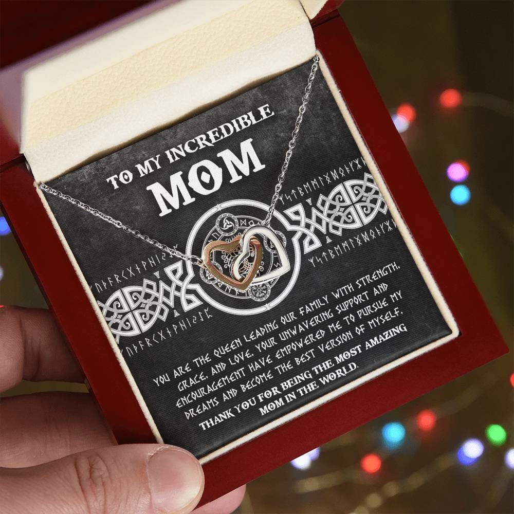 A person holds the To Mom, The Queen 2 - Interlocking Hearts Necklace adorned with cubic zirconia crystals in front of a card reading "To My Incredible Mom," while colorful, blurred lights shimmer in the background for a magical, personalized gift.