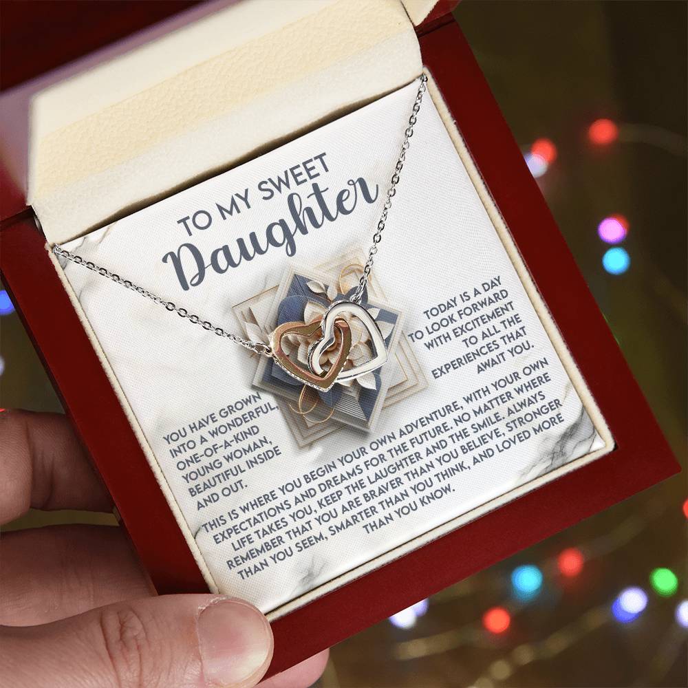 A hand presents an open red jewelry box with the Daughter-For The Future - Interlocking Hearts Necklace, made from surgical steel and sparkling cubic zirconia crystals. Shimmering colorful lights adorn the background, enhancing the heartfelt message inside for a daughter.