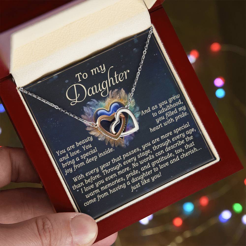 A hand holds an open jewelry box with a "Daughter-Like You - Interlocking Hearts Necklace" featuring shimmering cubic zirconia. The lid displays a heartfelt message to a daughter on a colorful background.