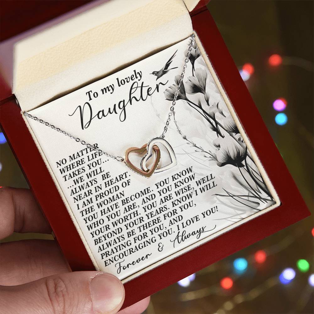 A hand presents an open red jewelry box, showcasing the Daughter-Heart Near - Interlocking Hearts Necklace decorated with cubic zirconia. A heartfelt message for a daughter is framed by floral designs in the background, with softly twinkling colorful lights.