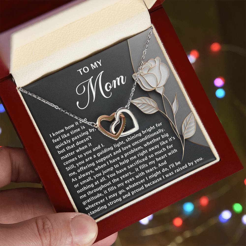 A person holds an open jewelry box featuring the Mom-Guiding Light - Interlocking Hearts Necklace in a rose gold finish, sparkling with cubic zirconia. A heartfelt "Mom" message on a rose-designed card is inside. The background softly twinkles with colorful lights.