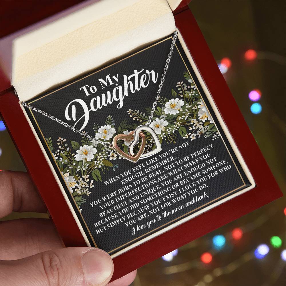 A close-up shows a hand holding an open red jewelry box with the Daughter-What You Do - Interlocking Hearts Necklace in rose gold. Inside the lid, a heartfelt "To My Daughter" letter features floral decorations, with blurred colored lights shimmering in the background.