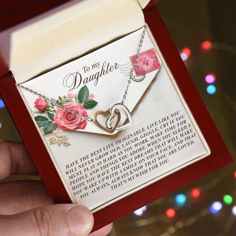 A hand presents a red jewelry box with the Daughter-My Wish - Interlocking Hearts Necklace. Within, pink roses frame a heartfelt message for a daughter. The necklace, crafted from surgical steel with rose gold finish and adorned with cubic zirconia crystals, shines against blurred colorful lights.